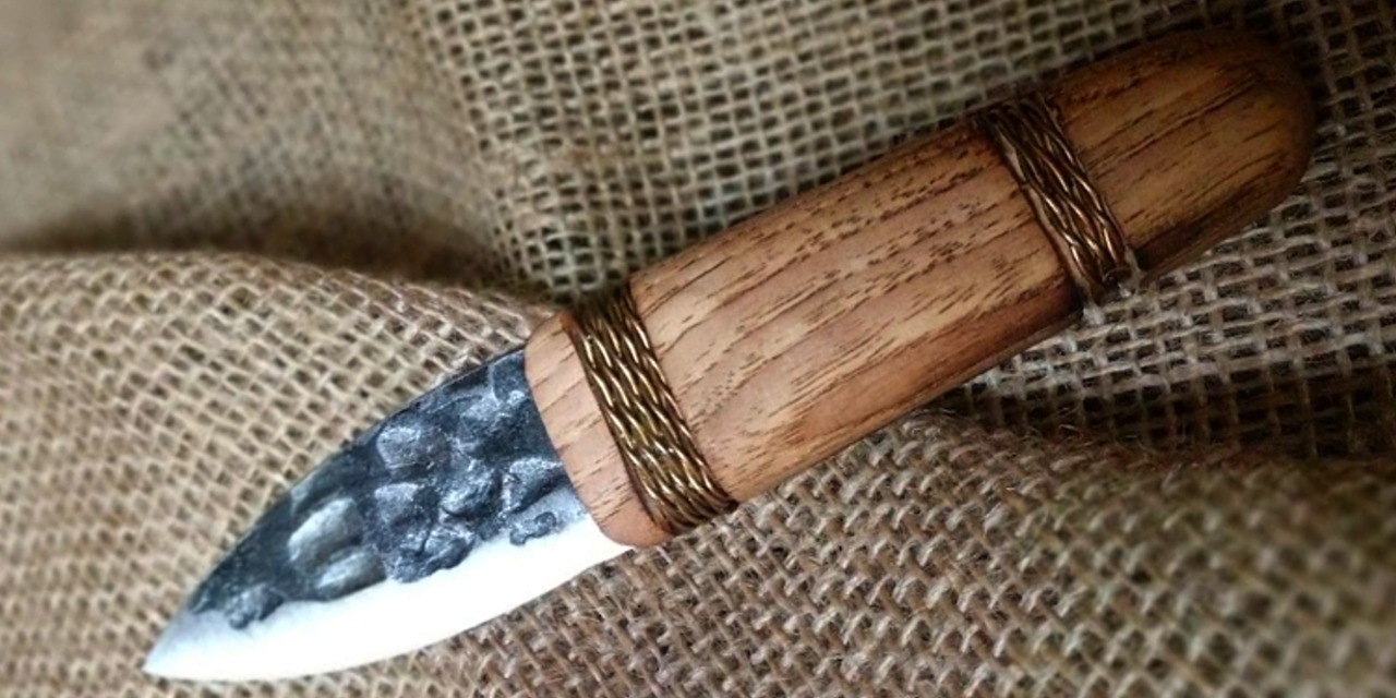 Primitive hammer forged steel fixed blade knife with a short arrowhead-shaped blade and wide wooden handle decorated with leather wraps at the top and bottom of the handle.