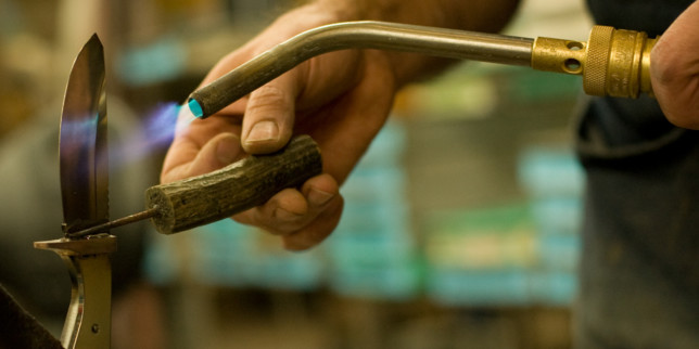 a hand holding a tool near a blade with a while hand tempering with a blowtorch
